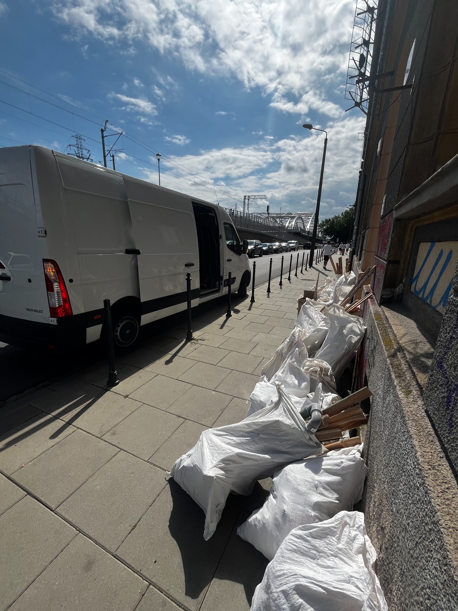 Biały van z otwartymi drzwiami zaparkowany na chodniku obok sterty białych worków z gruzem i fragmentami drewna, na tle miejskiego krajobrazu z wiaduktem kolejowym.