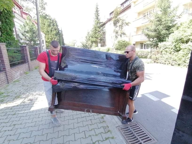 Dwóch mężczyzn w roboczych ubraniach przenosi owinięte folią pianino na tle chodnika i budynków.