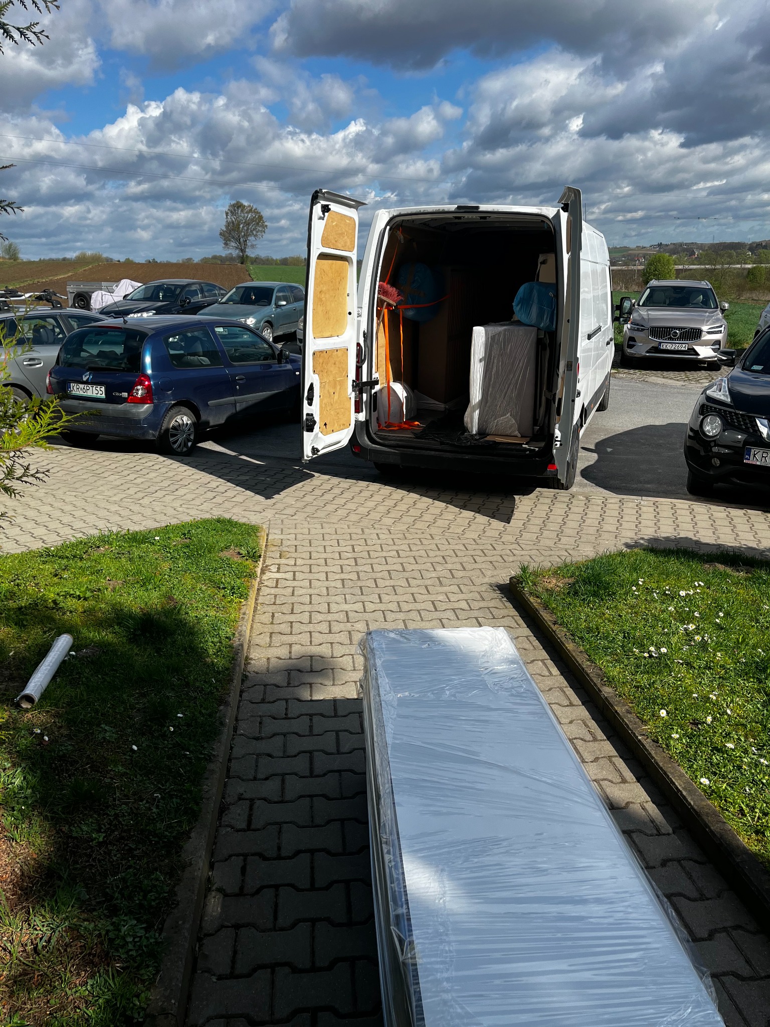 Biały furgon z otwartymi drzwiami bagażnika, częściowo załadowany meblami i sprzętem, stojący na brukowanym placu w słoneczny dzień. Na pierwszym planie spakowany w folię element, prawdopodobnie...