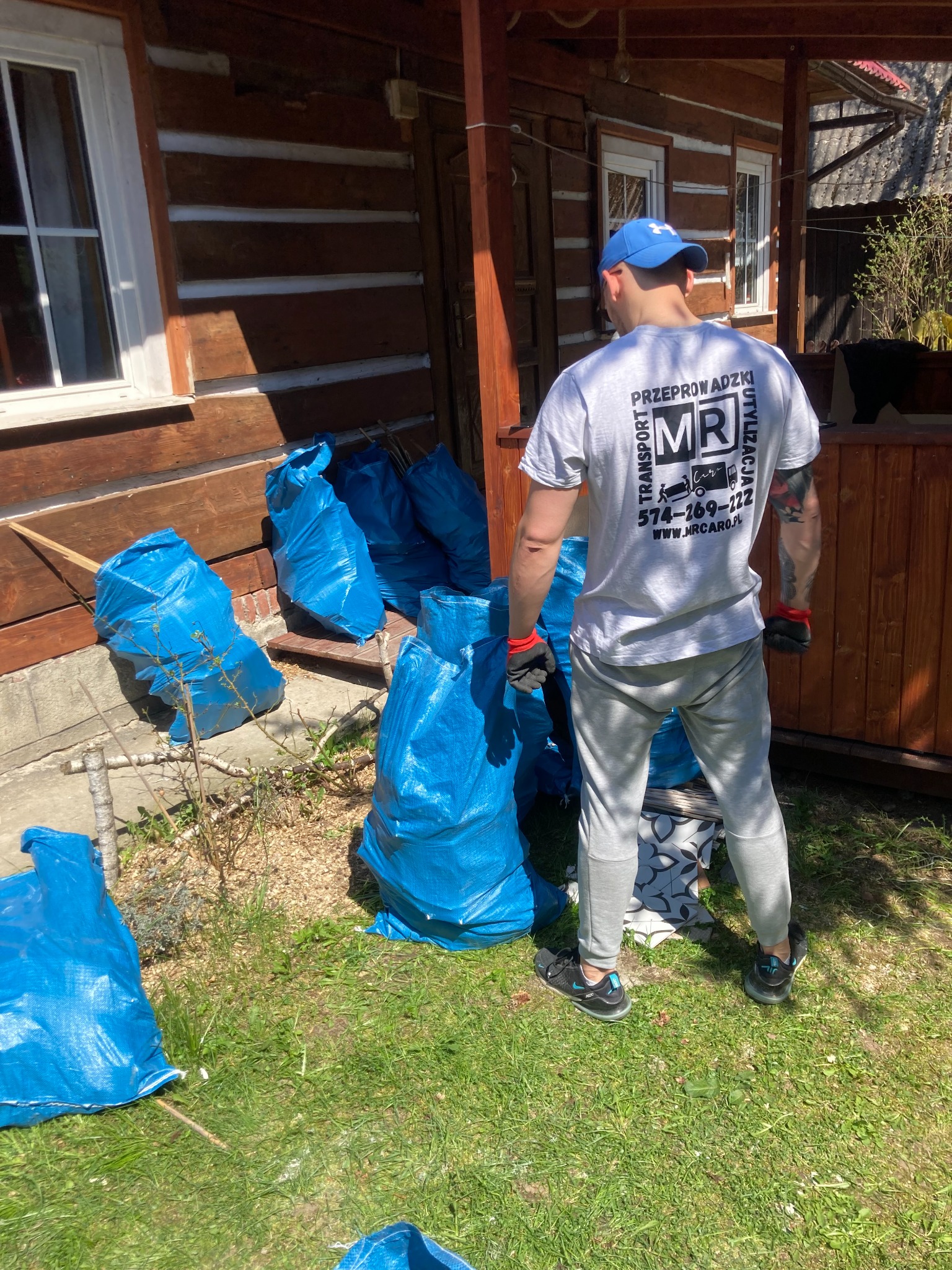 Mężczyzna w czapce i koszulce firmowej stoi tyłem do aparatu obok drewnianego domu, przed nim niebieskie worki wypełnione odpadami, prawdopodobnie po remoncie.