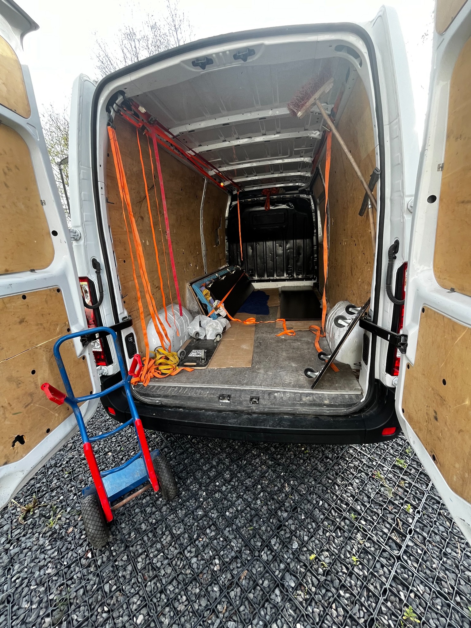 Wnętrze białego busa z otwartymi drzwiami, zabezpieczone pasami transportowymi, widoczny wózek transportowy na zewnątrz pojazdu, podłoże z kratki trawnikowej.