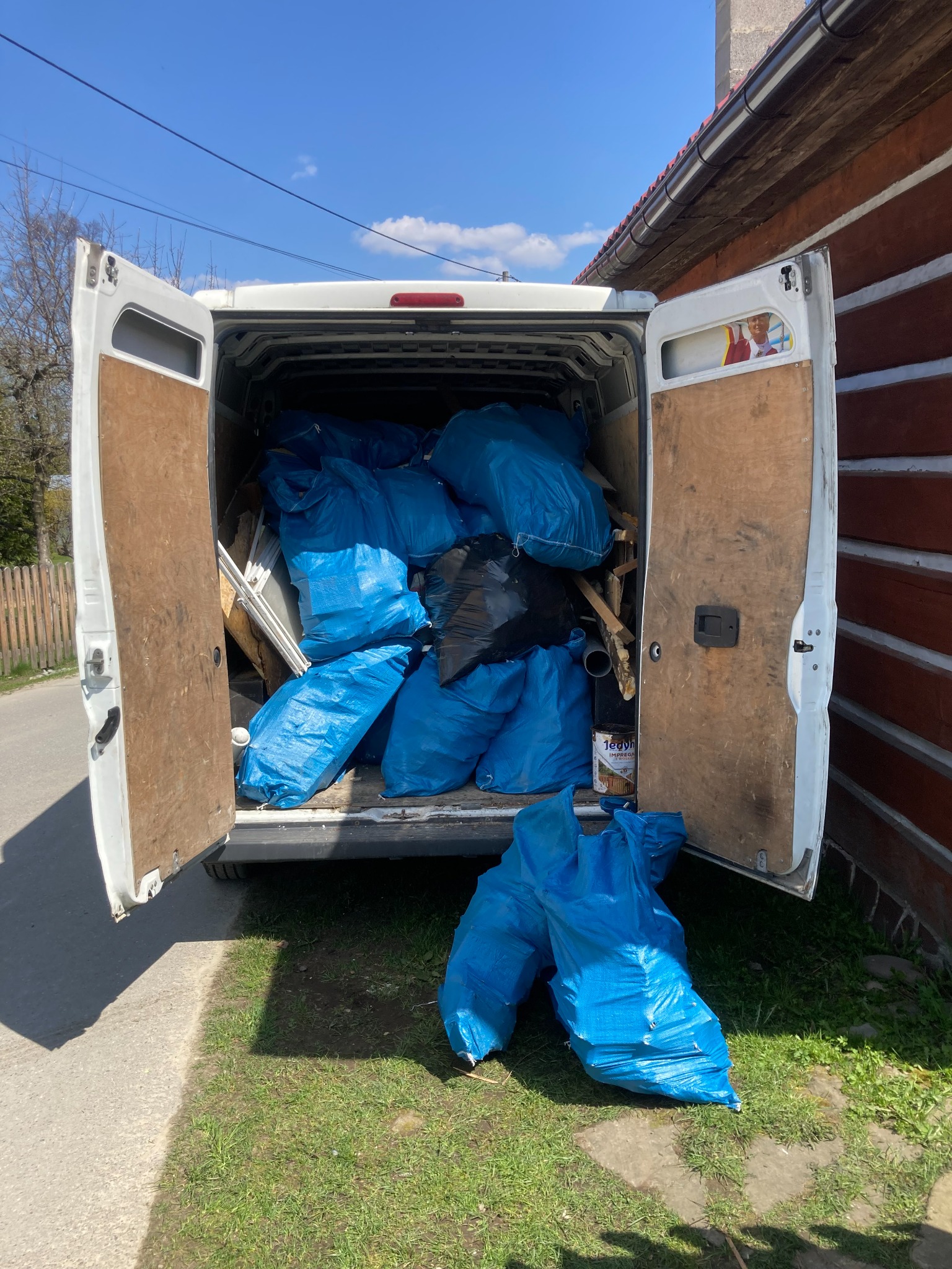 Otwarty bagażnik białego busa wypełniony niebieskimi workami z gruzem i odpadami budowlanymi, na zewnątrz dwa dodatkowe worki, widoczny fragment drewnianego domu w tle.