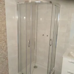 Narożna kabina prysznicowa z przezroczystego szkła z chromowanymi uchwytami w łazience wyłożonej jasnymi płytkami, widoczna mozaika na ścianie i biała szafka z umywalką.