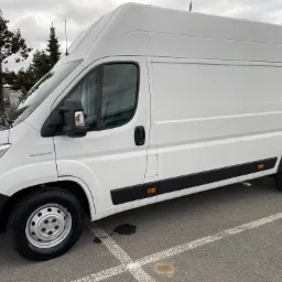 Biały samochód dostawczy Fiat Ducato z wysokim dachem, zaparkowany na parkingu z widocznymi plamami na asfalcie i drzewami w tle.