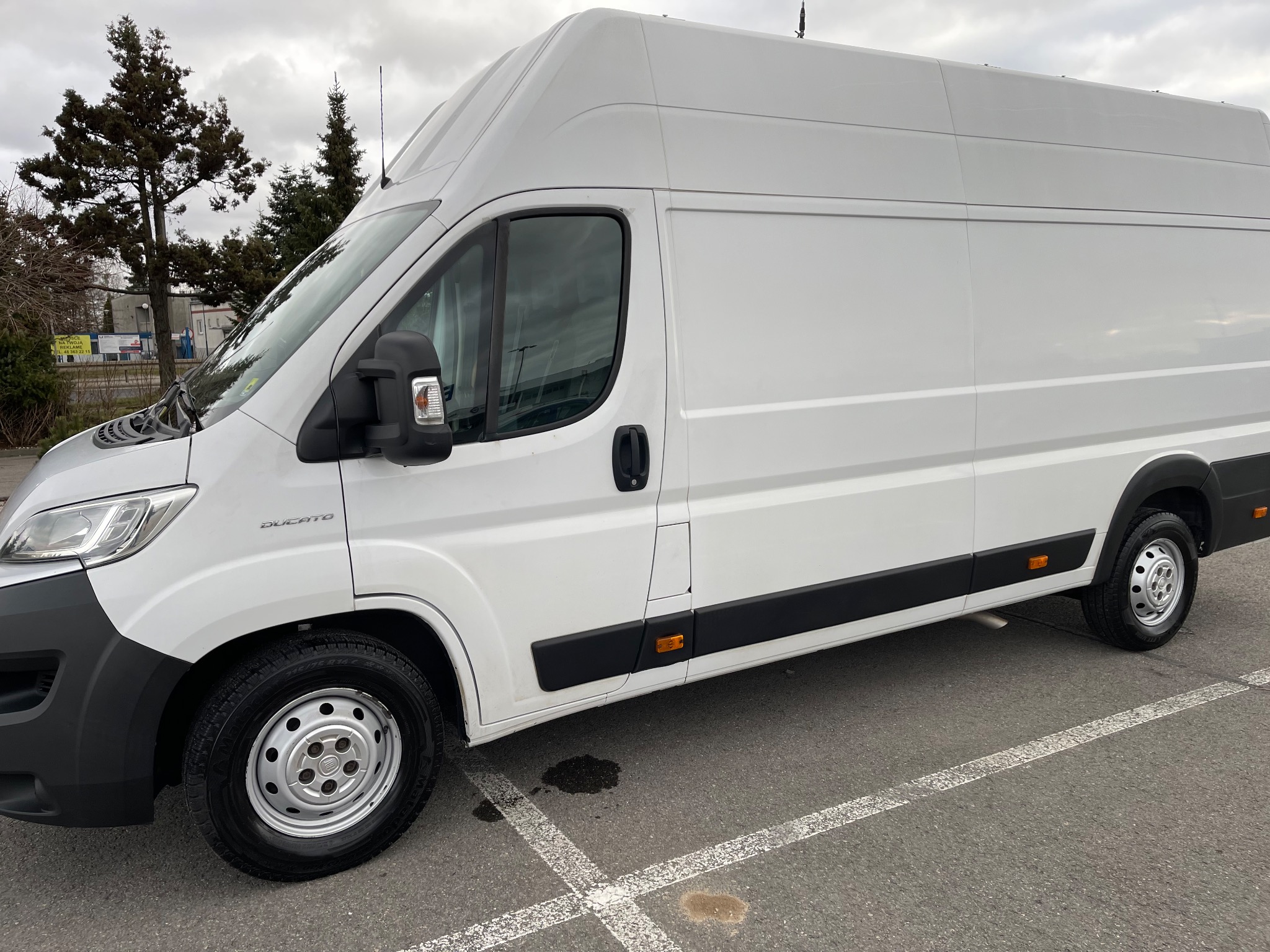 Biały samochód dostawczy Fiat Ducato z wysokim dachem, zaparkowany na parkingu z widocznymi plamami na asfalcie i drzewami w tle.