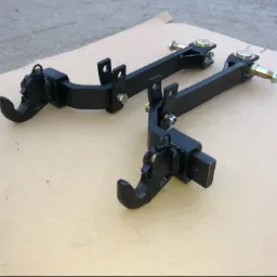 Dwa czarne, metalowe zaczepy z elementami obrotowymi i śrubami, leżące na kartonowym podłożu, prawdopodobnie elementy do maszyn rolniczych.