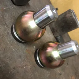 Dwa metalowe przeguby kulowe, jeden z otworem, leżące na szorstkiej, metalowej powierzchni roboczej obok młotka.