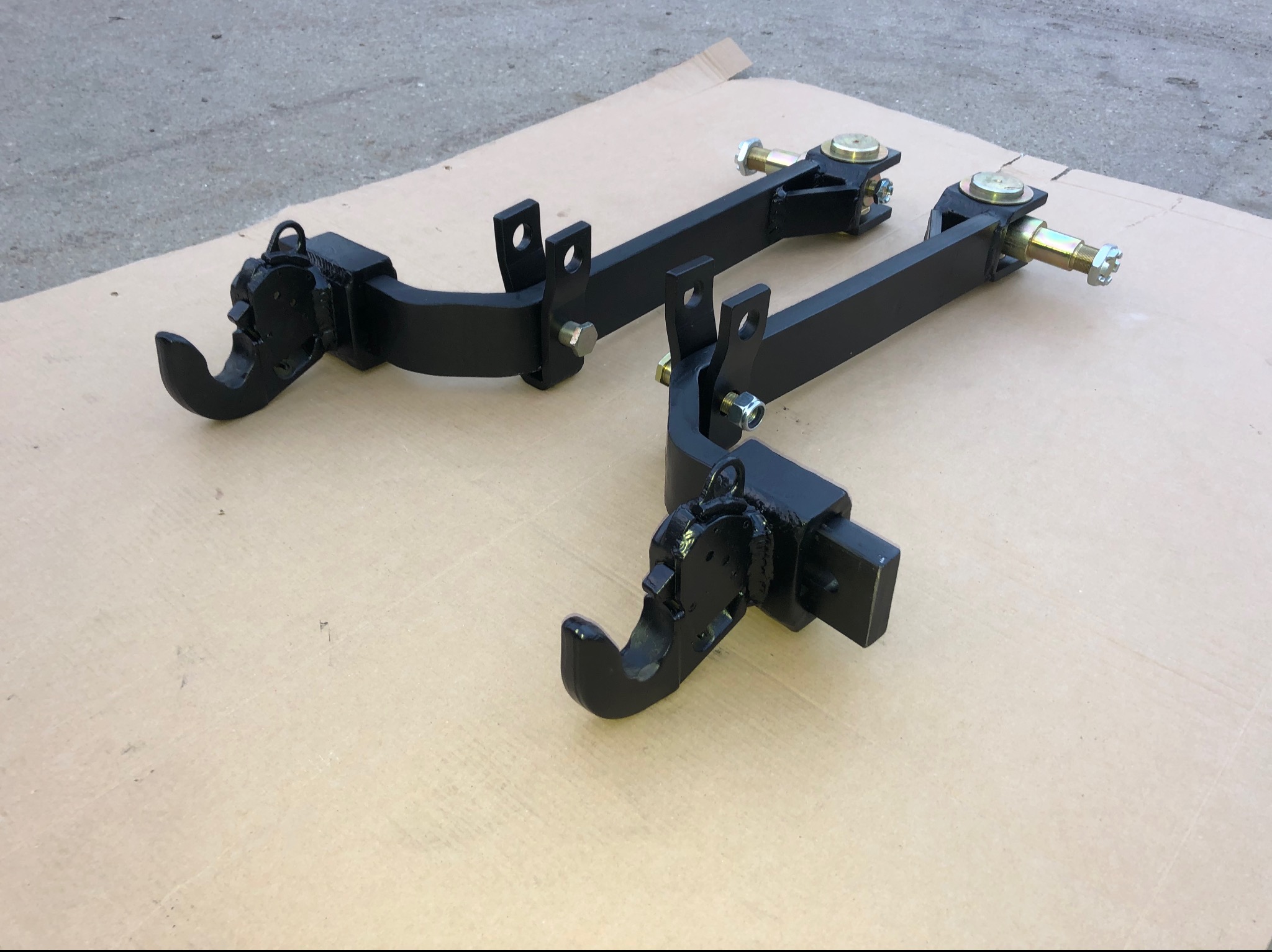 Dwa czarne, metalowe zaczepy z elementami obrotowymi i śrubami, leżące na kartonowym podłożu, prawdopodobnie elementy do maszyn rolniczych.