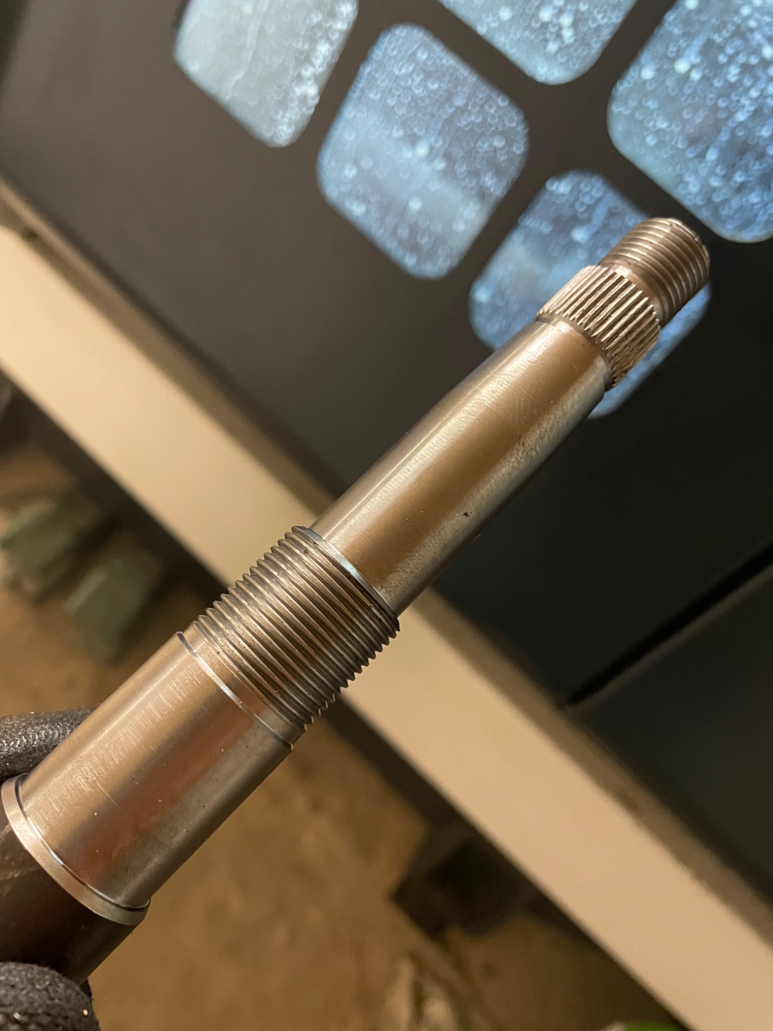 Metalowy wałek z gwintowanymi sekcjami, trzymany w rękawicy roboczej na tle monitora z rozmytym obrazem.