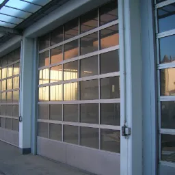 Zewnętrzny widok na trzy segmentowe bramy garażowe z przeszklonymi panelami w szarej ramie, z widocznymi odbiciami budynków w szybach.