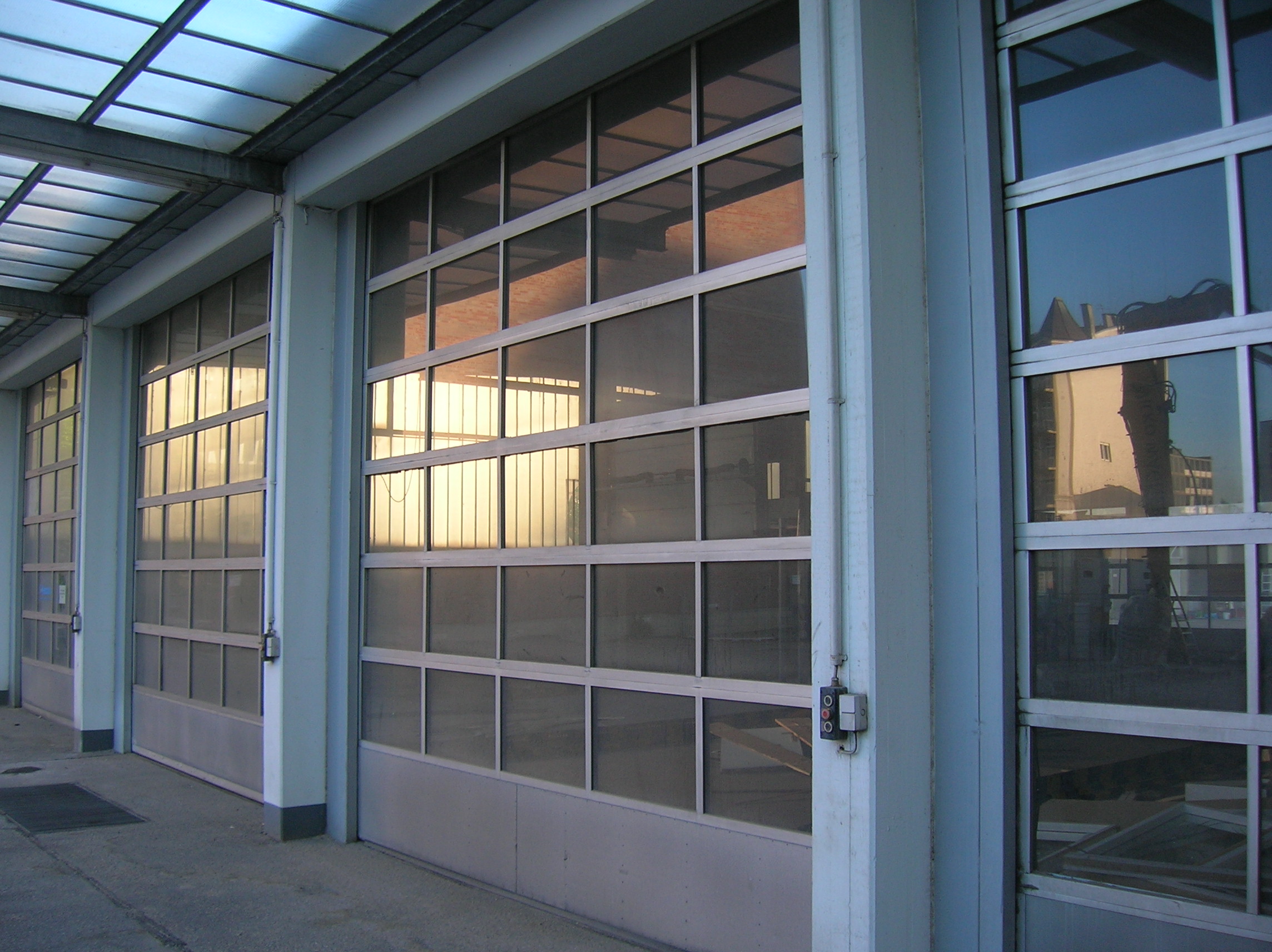 Zewnętrzny widok na trzy segmentowe bramy garażowe z przeszklonymi panelami w szarej ramie, z widocznymi odbiciami budynków w szybach.