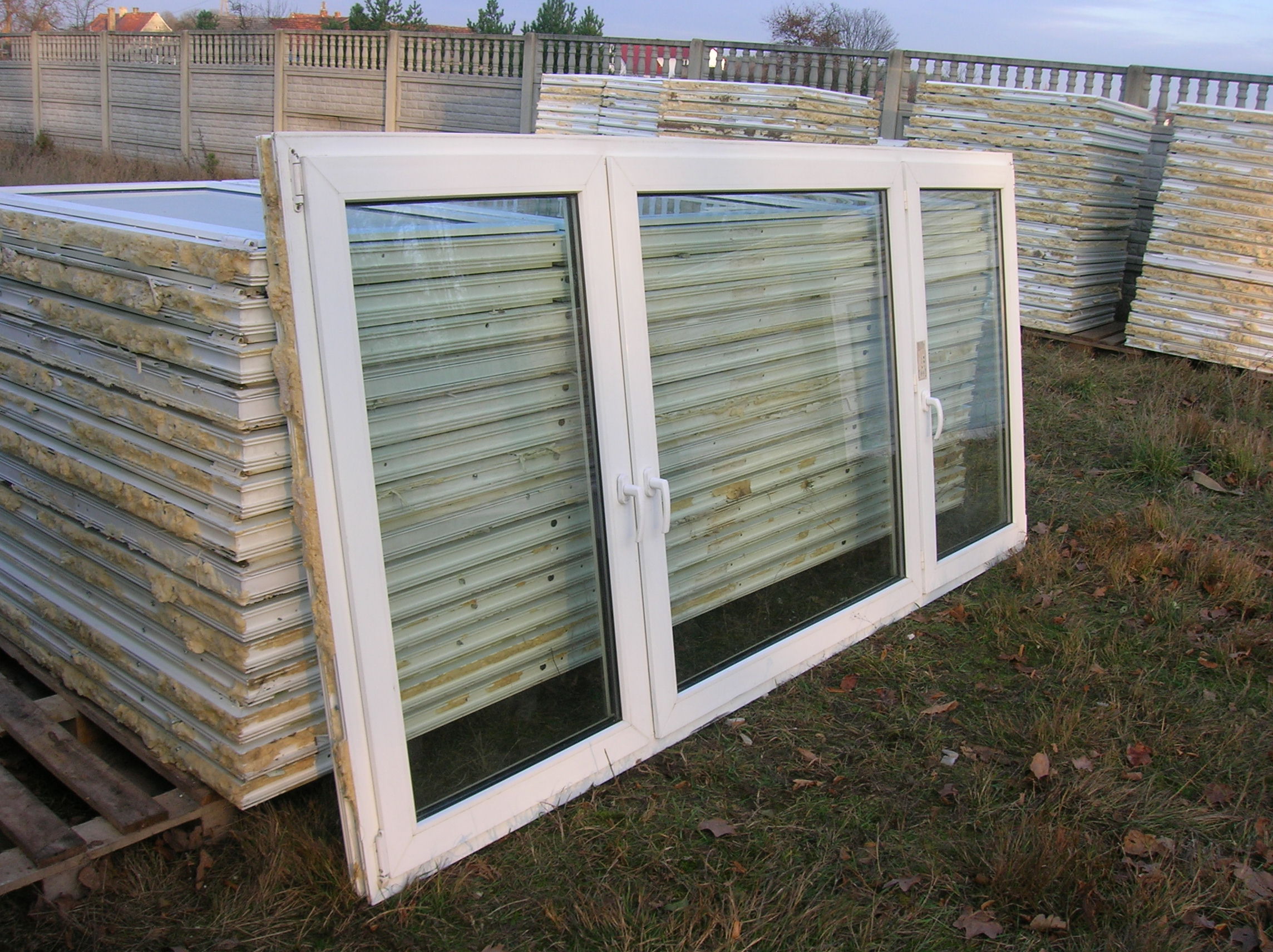 Białe, trzyskrzydłowe okno PCV oparte o stos płyt warstwowych z widocznym rdzeniem izolacyjnym, składowane na palecie, na tle betonowego ogrodzenia.