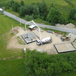 F.U.P.H. TRANS-BET STANISŁAW RAPACZ - Z góry: wylewanie betonu do fundamentów domu z użyciem pompy, betonomieszarka na budowie, teren zielony i droga w tle. Widoczny postęp prac budowlanych.