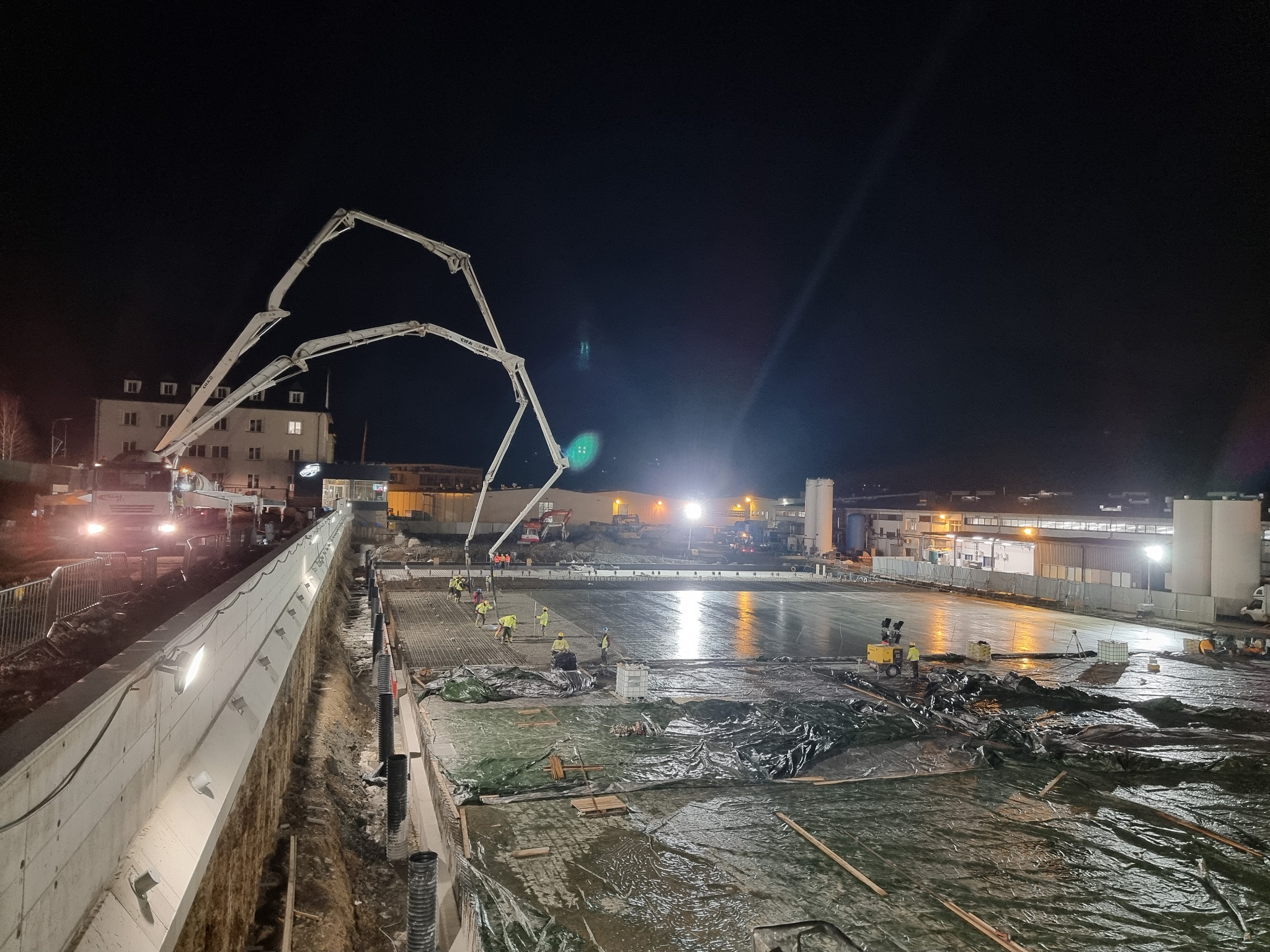Nocne wylewanie betonu z pompą na budowie; widoczni pracownicy w żółtych kamizelkach, zbrojenie i folia. Oświetlony teren budowy.