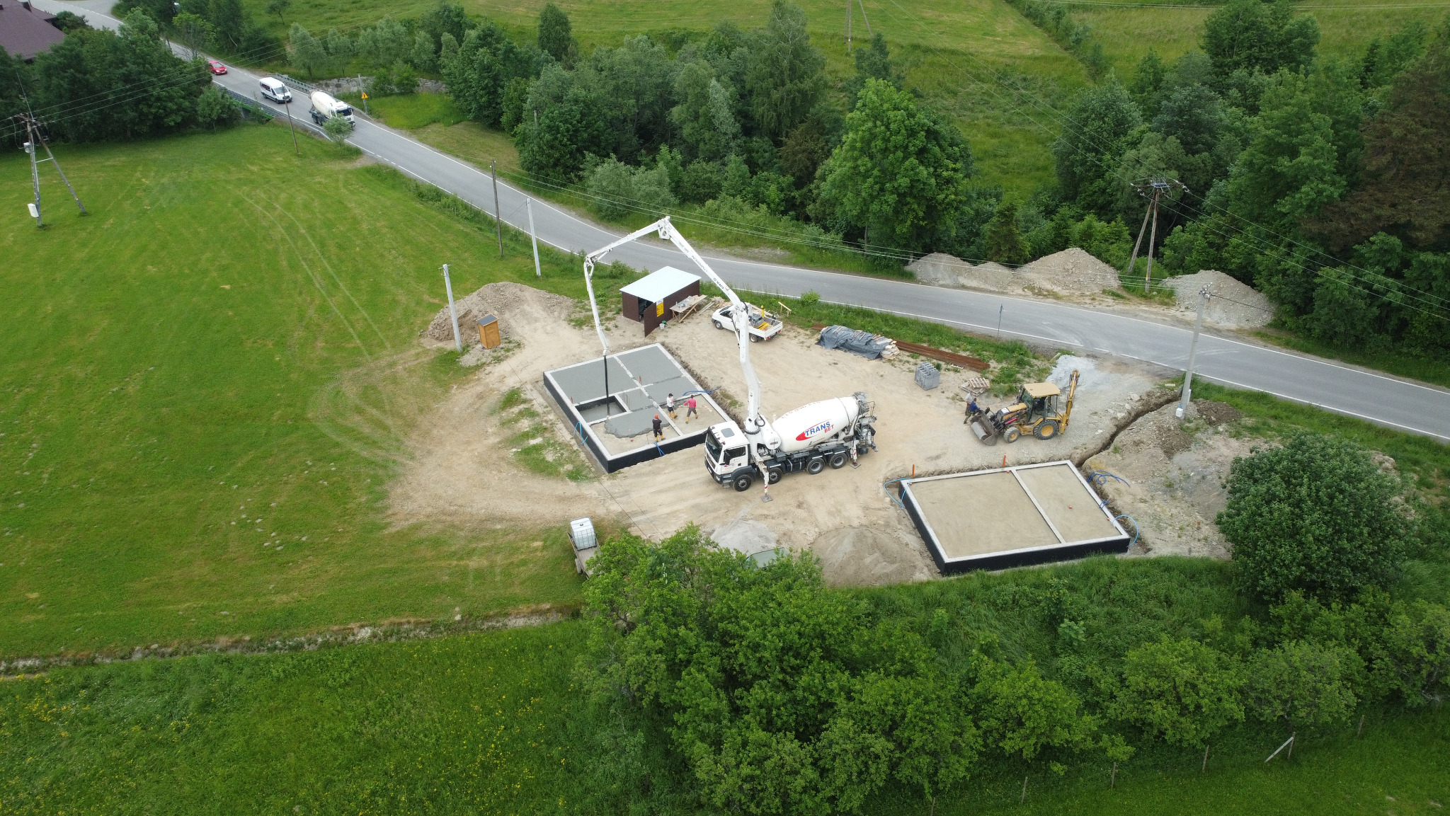 Z góry: wylewanie betonu do fundamentów domu z użyciem pompy, betonomieszarka na budowie, teren zielony i droga w tle. Widoczny postęp prac budowlanych.