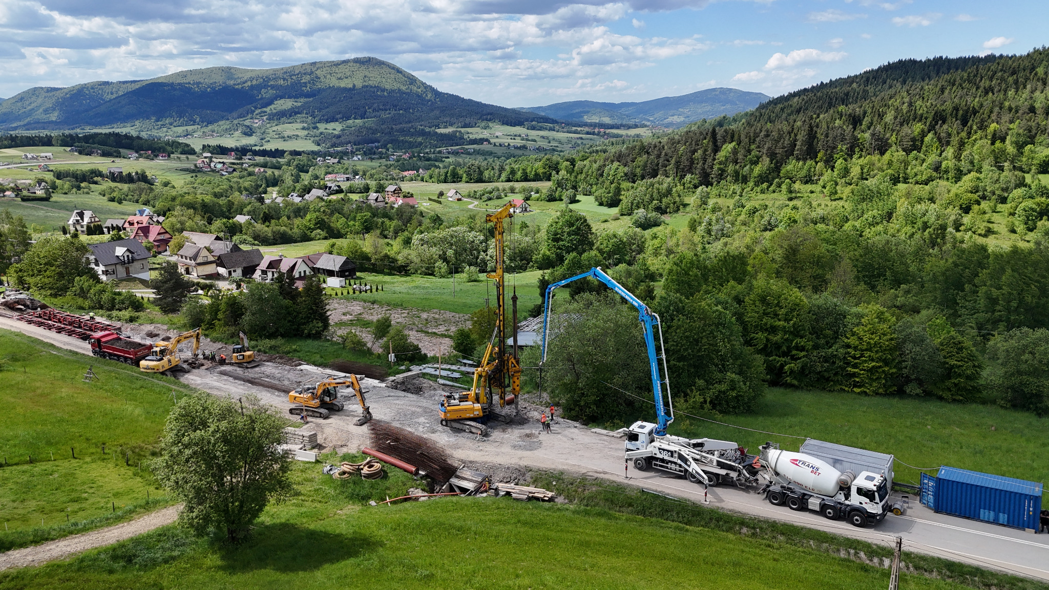 Szeroki kadr z góry: maszyny budowlane (koparki, wiertnica, betonomieszarka z pompą) pracują na tle malowniczego krajobrazu górskiego i zabudowań.