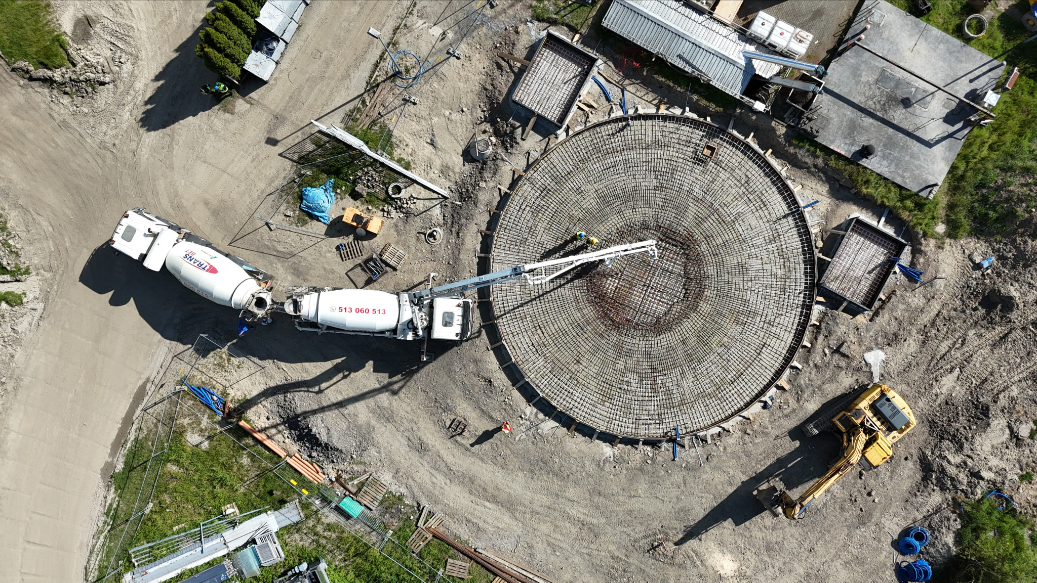 Z góry: betonowóz z pompą w akcji przy okrągłym zbrojeniu fundamentu. Obok koparka i przygotowane szalunki. Widok na plac budowy z drona.