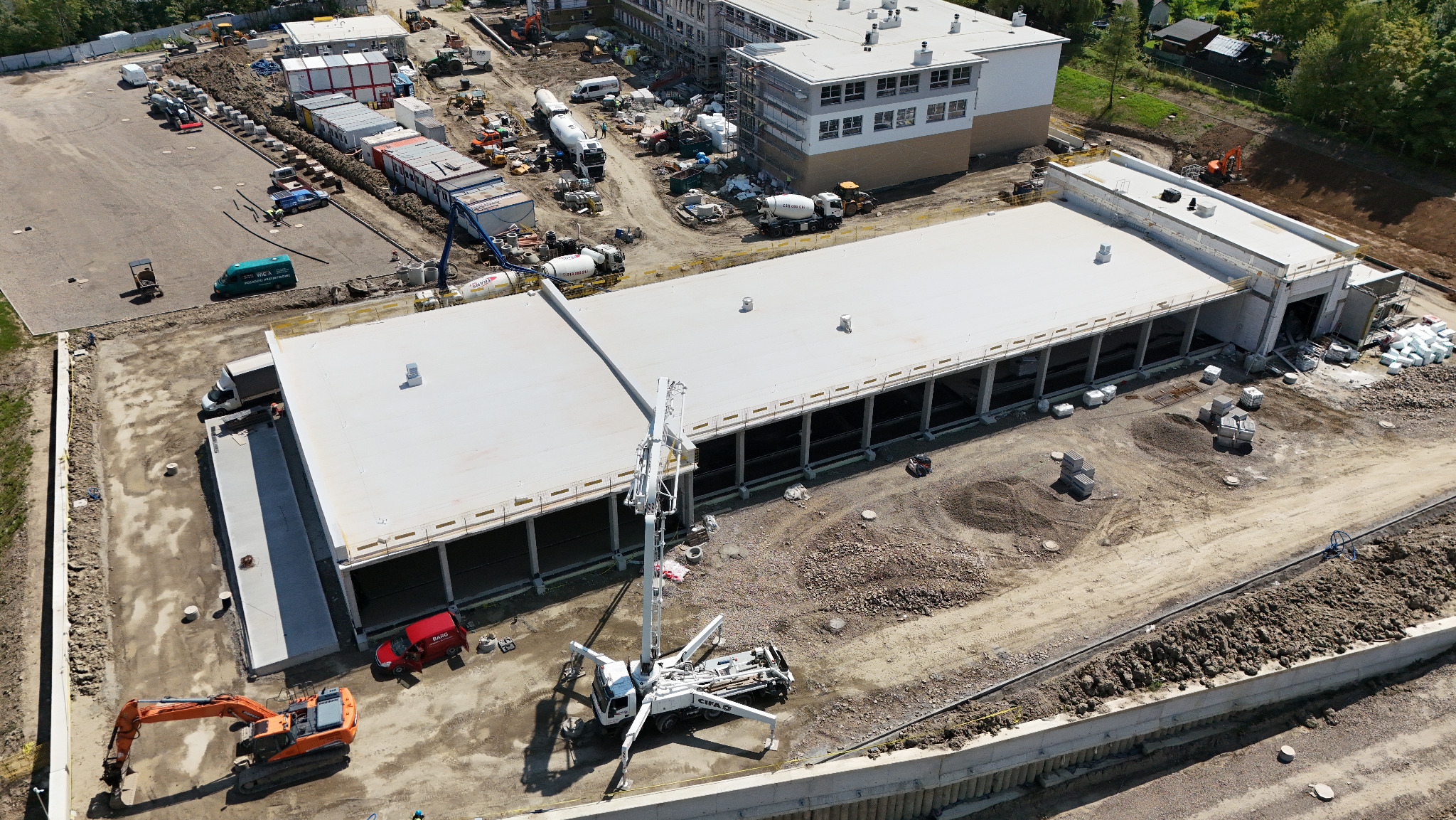 Betonowanie dachu budynku przemysłowego z użyciem pompy do betonu. Widoczne miksokrety i prace ziemne z użyciem koparki. Ujęcie z drona, wysoka perspektywa.