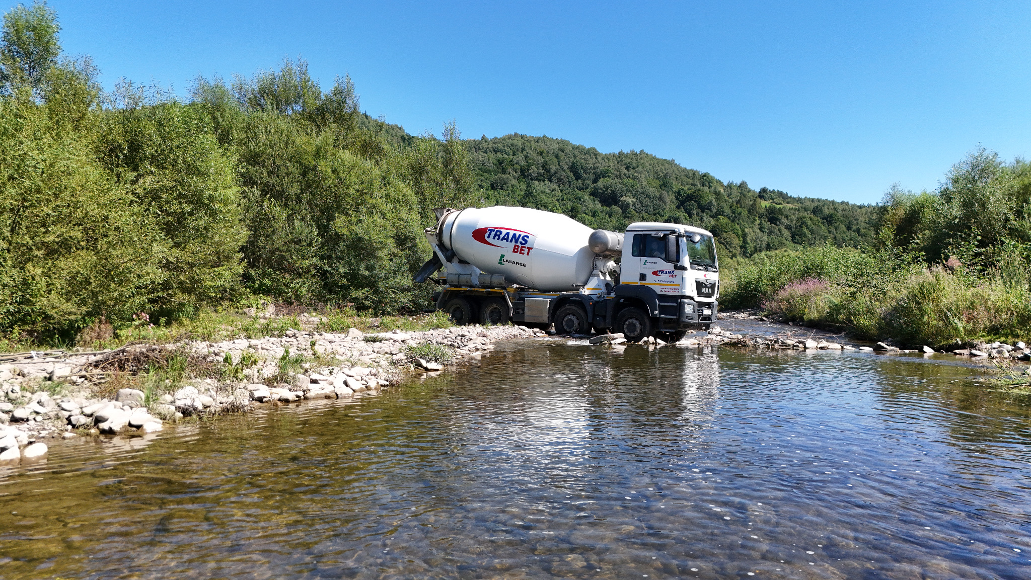 Ciężarówka z betonomieszarką marki MAN przejeżdża przez płytki strumień na tle zieleni, w pogodny dzień.