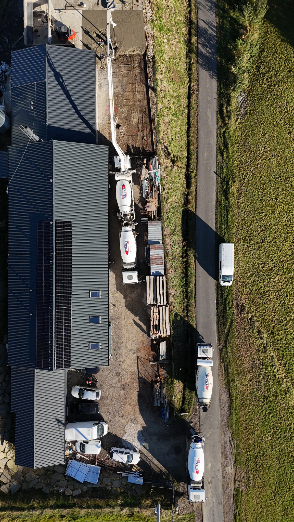 Widok z góry na plac budowy z betoniarkami, pompą do betonu i wylewką fundamentów. Widoczne panele słoneczne na dachu budynku i zaparkowane auta.