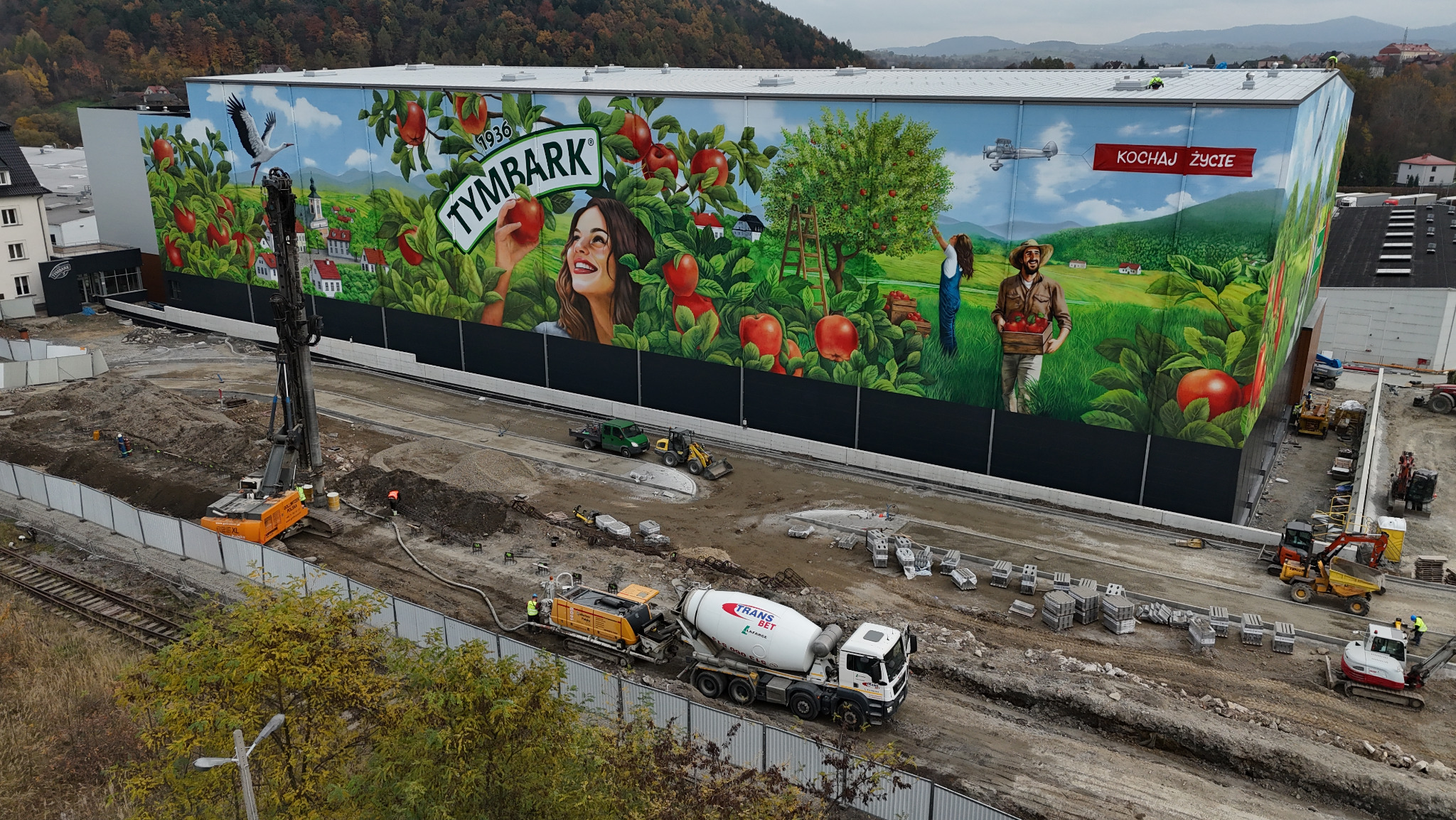 Betoniarka Trans-Bet Limanowa w trakcie prac na placu budowy, na tle budynku z muralem Tymbarku. Widoczny ciężki sprzęt i materiały budowlane.
