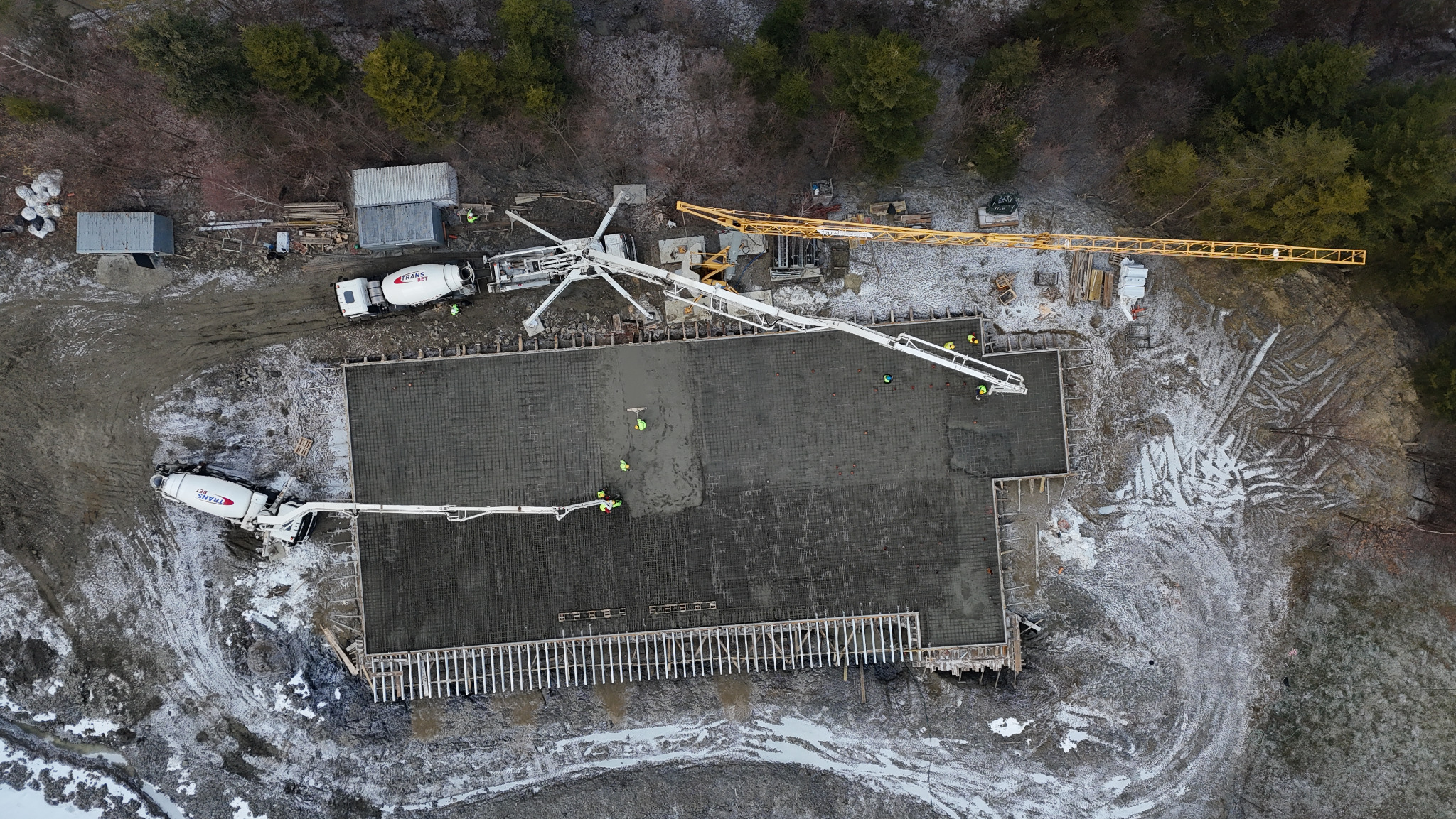 Z góry: Wylewanie fundamentów z betonomieszarek na zbrojenie, widoczny żuraw i pracownicy w żółtych kamizelkach. Częściowo ośnieżony teren budowy.