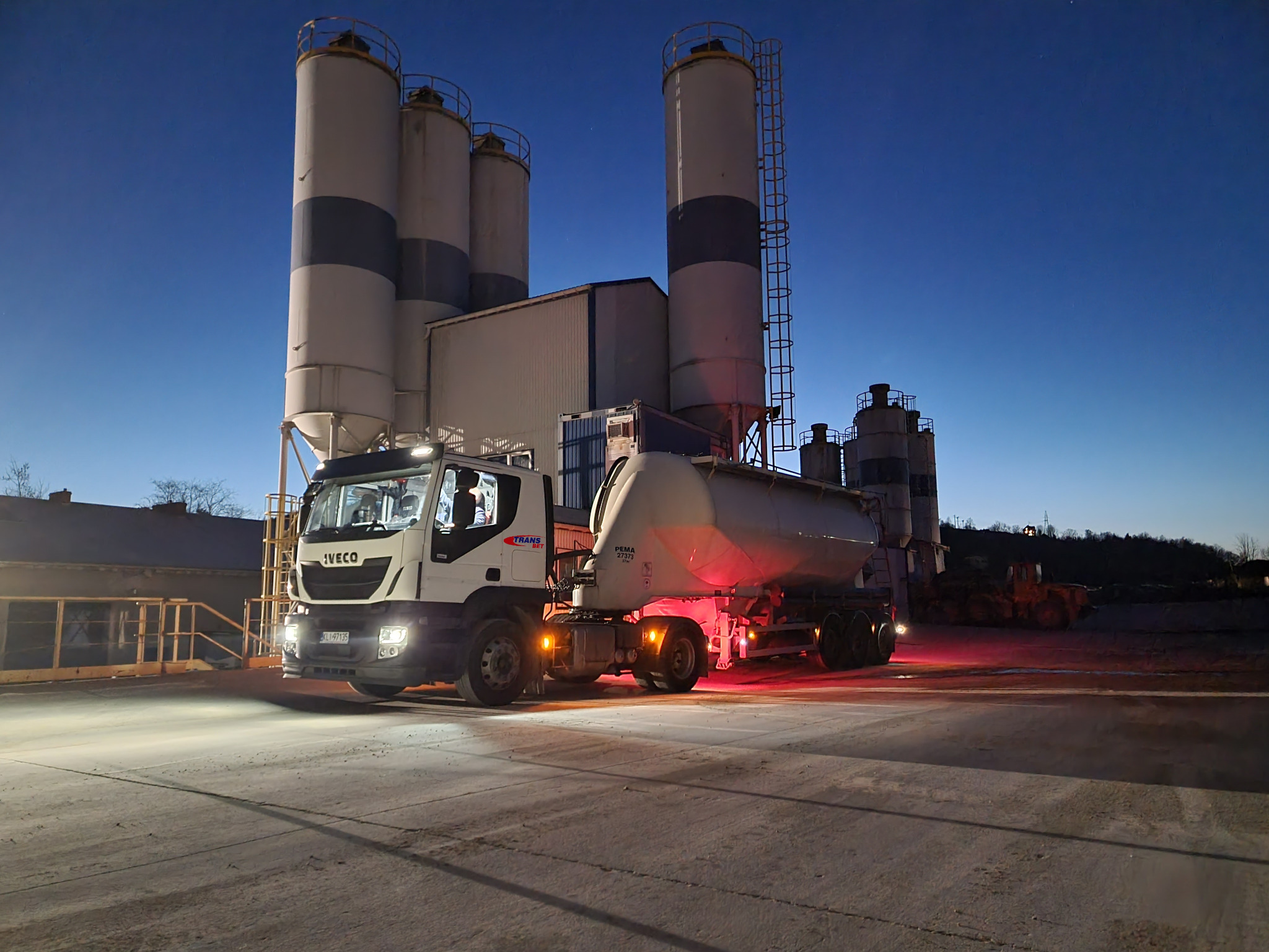 Ciężarówka Iveco z naczepą cysterną podświetlona światłami, na tle silosów do przechowywania cementu i betonu, nocna sceneria.
