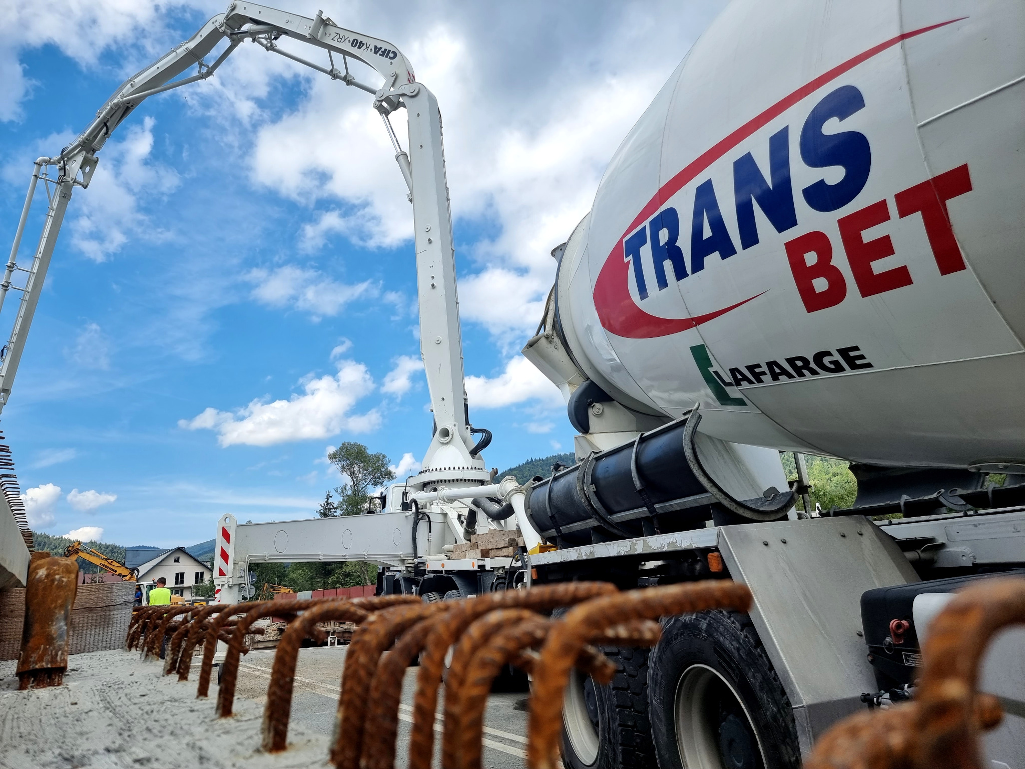 Ciężarówka z betonomieszarką marki Trans Bet Lafarge z wysięgnikiem pompy do betonu na budowie, widok zza zbrojenia.