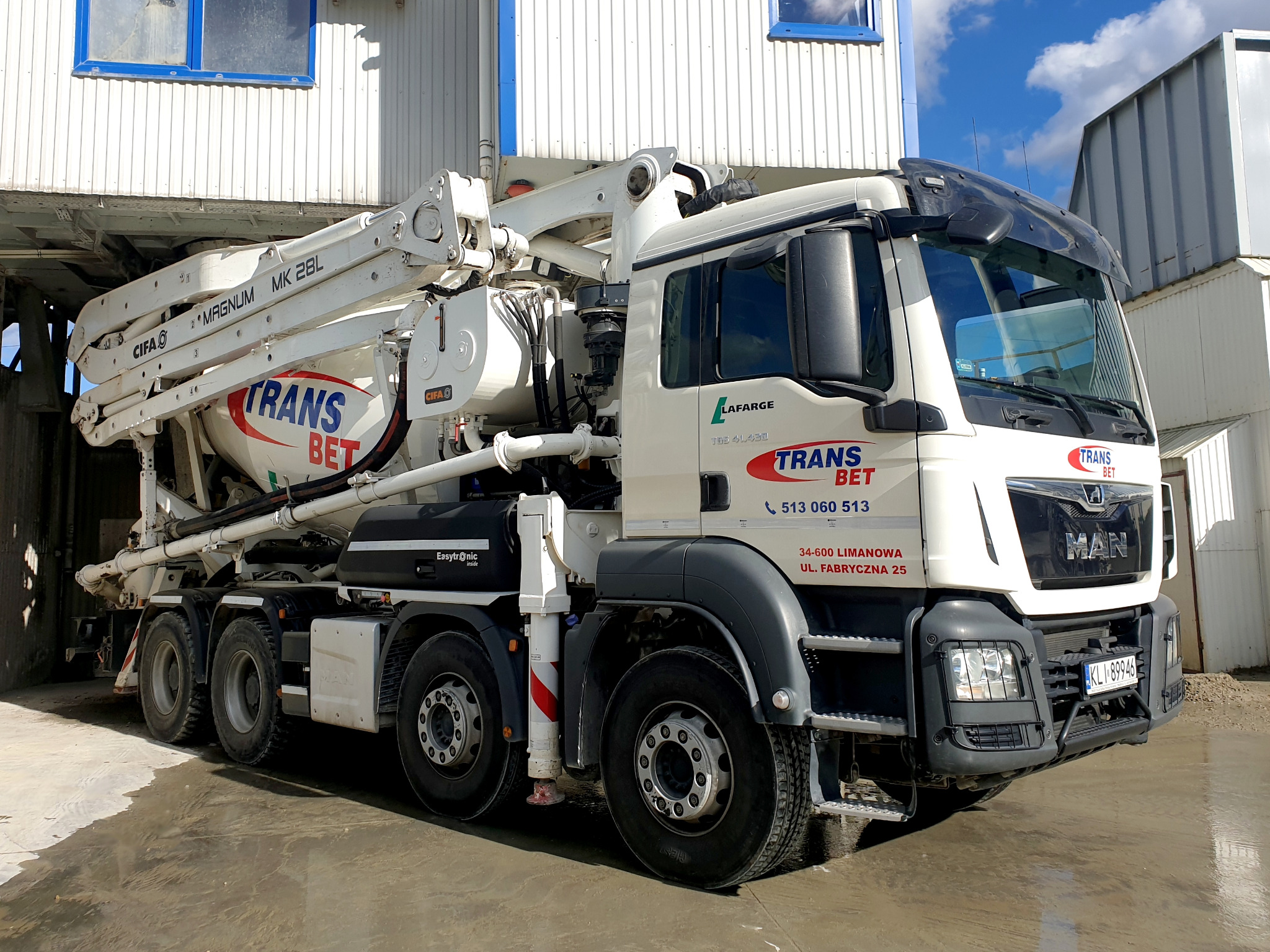 Ciężarówka MAN z pompą do betonu CIFA Magnum MK 28L, z logo Trans Bet, zaparkowana na terenie betoniarni, widok z przodu na tle budynku z blachy falistej i nieba.