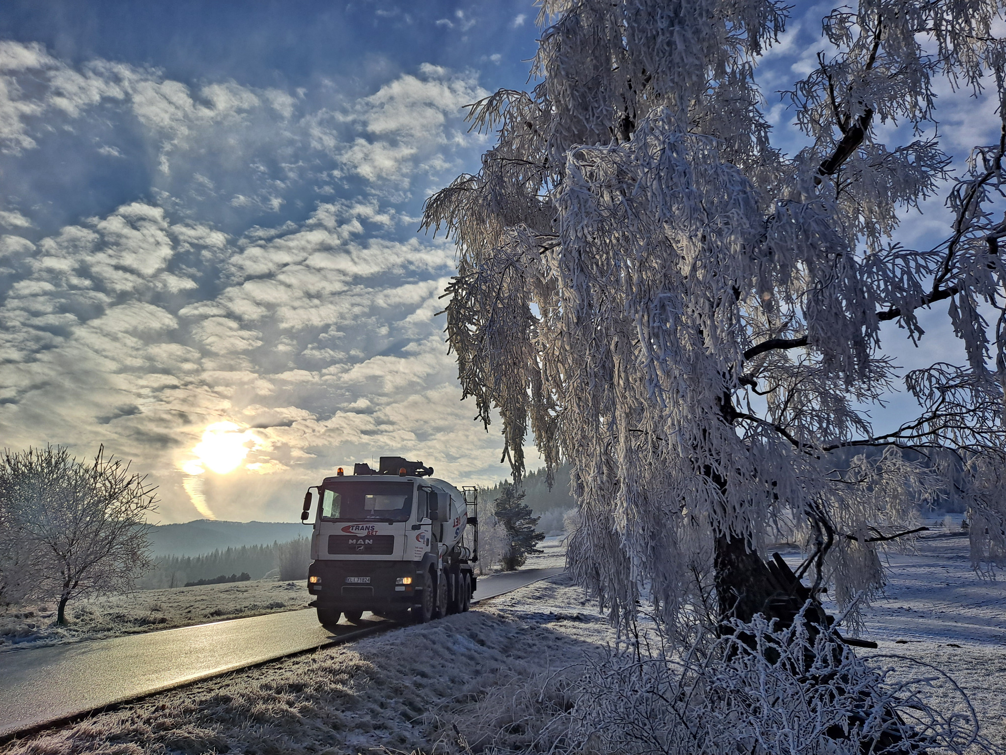 Betonomieszarka MAN w zimowym krajobrazie, ośnieżone drzewa i droga, słońce przebija się przez chmury.