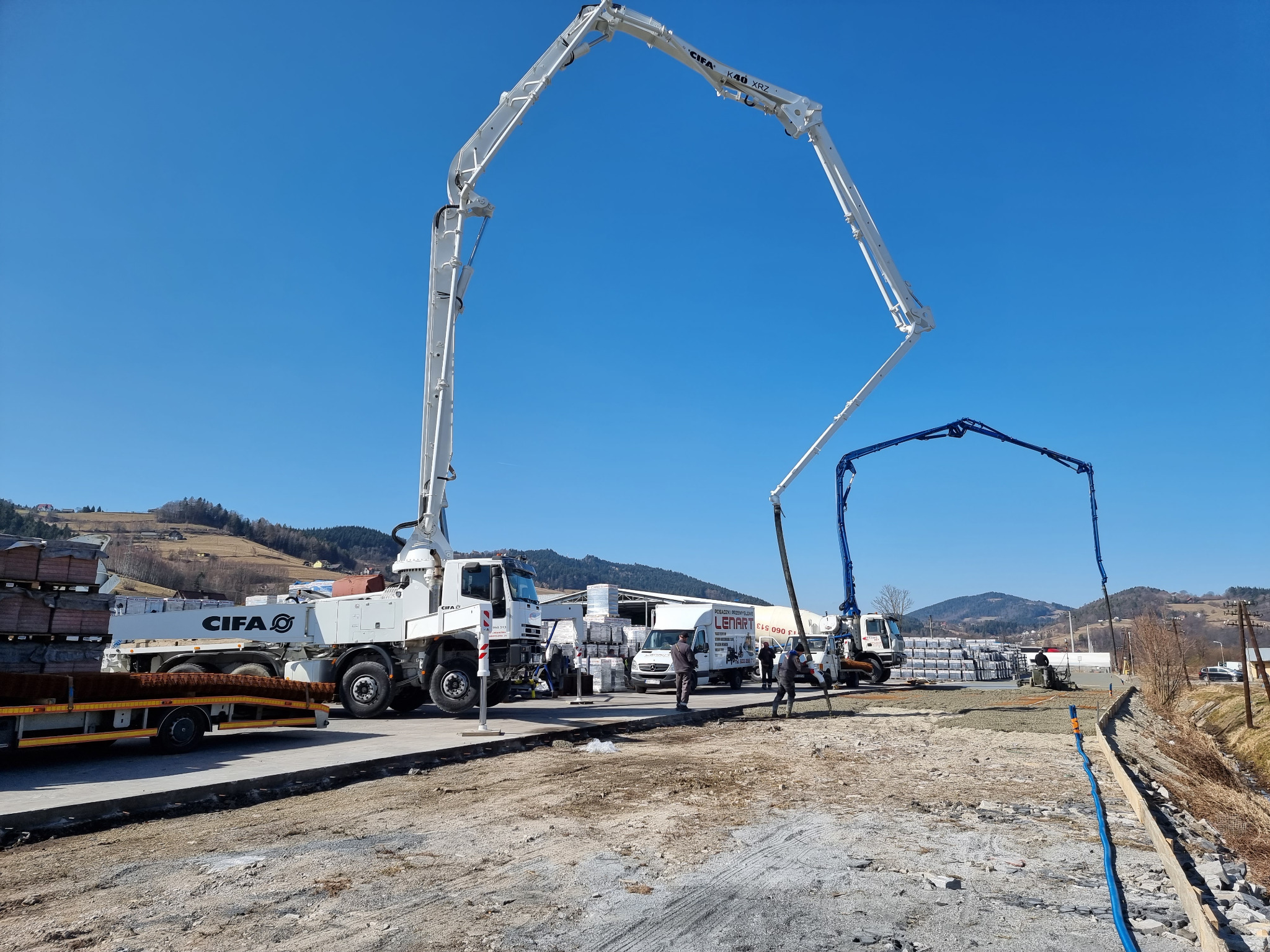 Dwie pompy do betonu (biała CIFA K40 XRZ i niebieska) na placu budowy, w tle palety z cegłami, teren górzysty i błękitne niebo.
