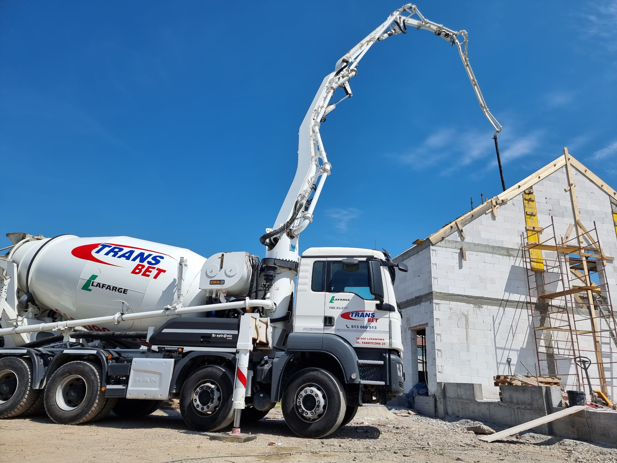 Ciężarówka z pompą do betonu marki MAN w trakcie wylewania betonu na budowę domu z białych bloczków, widoczne rusztowanie.