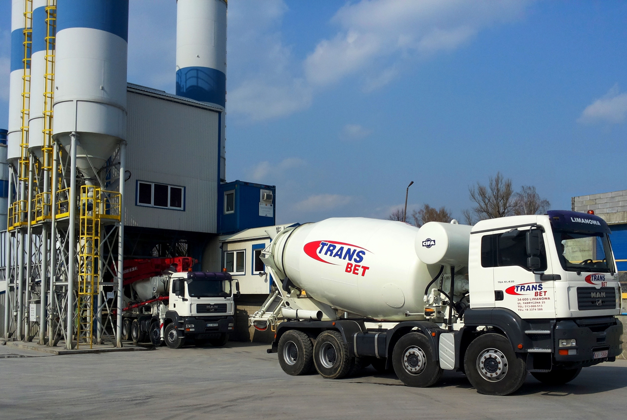 Dwa samochody betonomieszarki z logo Trans-Bet na tle silosów betoniarni w słoneczny dzień.