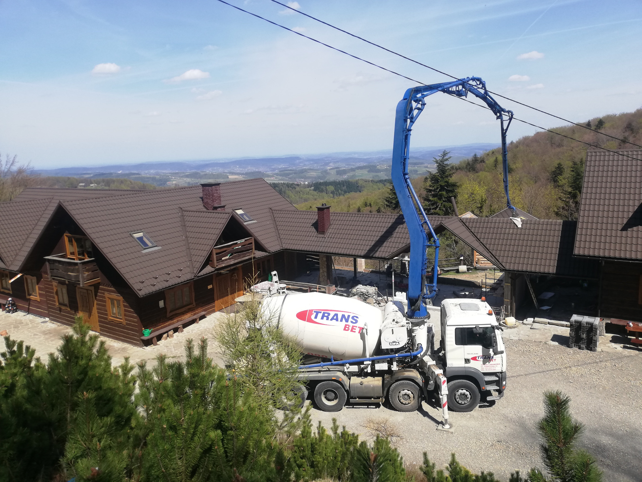 Betonomieszarka z wysięgnikiem pompy stoi przed drewnianym domem na tle górskiego krajobrazu w słoneczny dzień.