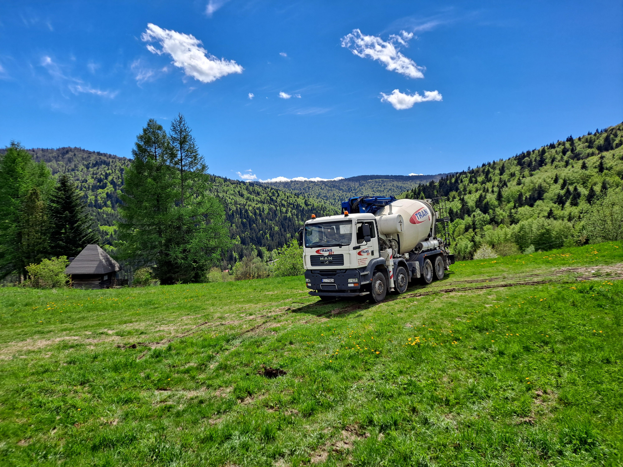 Ciężarówka do transportu betonu marki MAN z napisem TRANS BET na tle malowniczego krajobrazu z zielonymi wzgórzami, błękitnym niebem i drewnianą chatą w oddali.