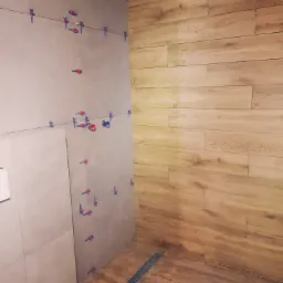 Ściana łazienki w trakcie układania płytek, widoczne klipsy poziomujące, oraz ułożona drewnopodobna podłoga z odpływem liniowym.