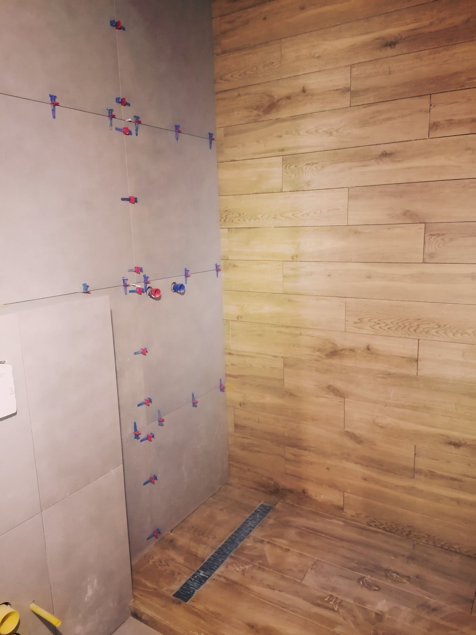 Ściana łazienki w trakcie układania płytek, widoczne klipsy poziomujące, oraz ułożona drewnopodobna podłoga z odpływem liniowym.