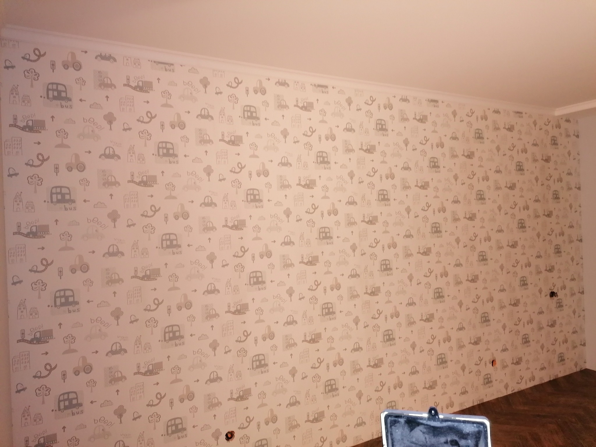 Ściana w pokoju dziecięcym pokryta jasną tapetą z szarymi wzorami pojazdów, drzew i budynków, widoczne gniazdka elektryczne i fragment podłogi z paneli.