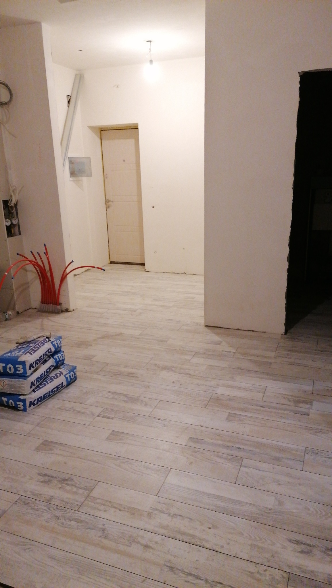 Ułożona podłoga z płytek imitujących drewno w trakcie remontu mieszkania, widoczne worki z klejem i rury instalacji grzewczej przy ścianie.