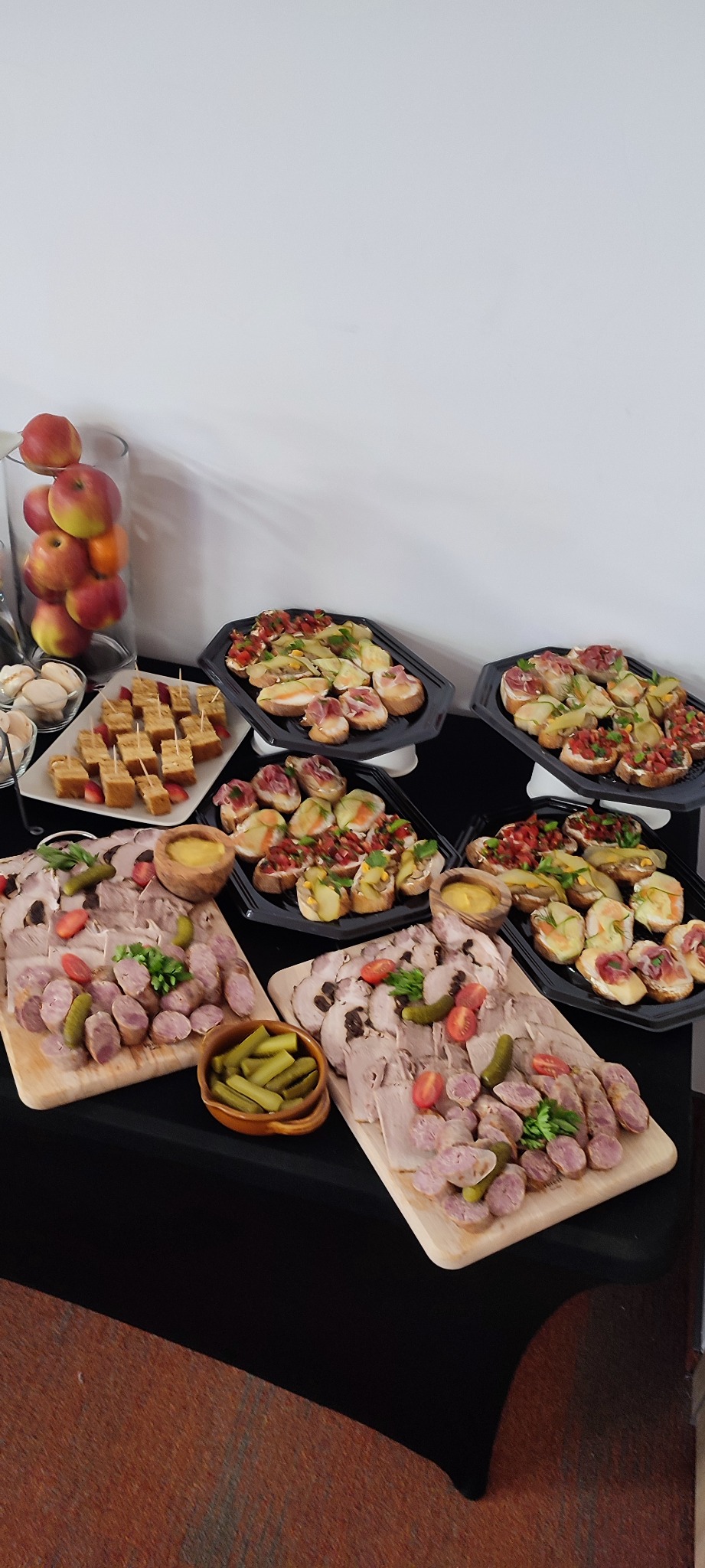 Stół cateringowy z przekąskami: kanapki na czarnych tacach, deski z wędlinami i ogórkami konserwowymi, miseczka z dipem, jabłka w szklanym wazonie i małe ciasteczka z truskawkami.
