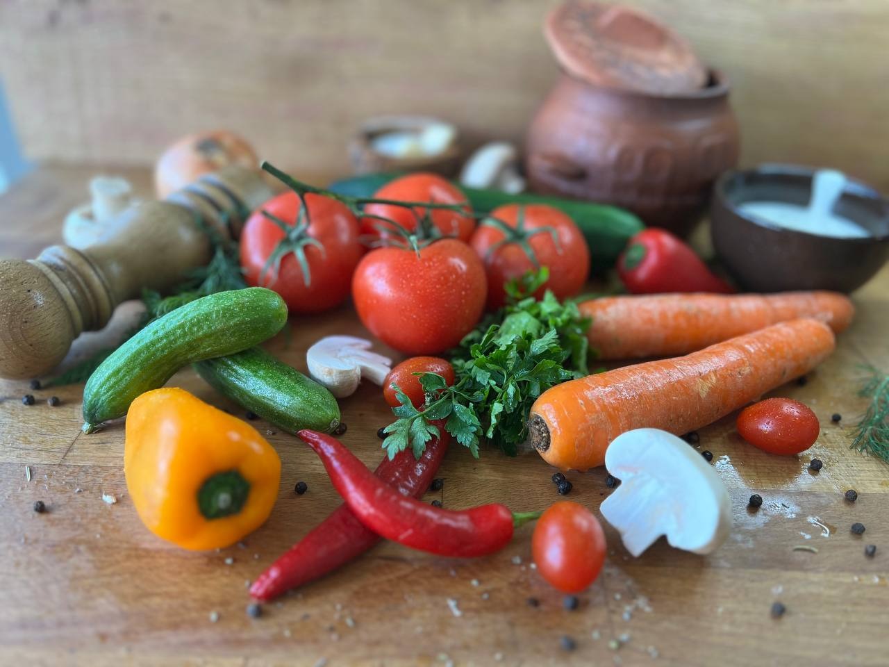 Kompozycja świeżych warzyw na drewnianej desce: pomidory na gałązce, ogórki, marchew, papryka żółta i czerwona, grzyby, cebula, przyprawy, gliniane naczynia w tle.