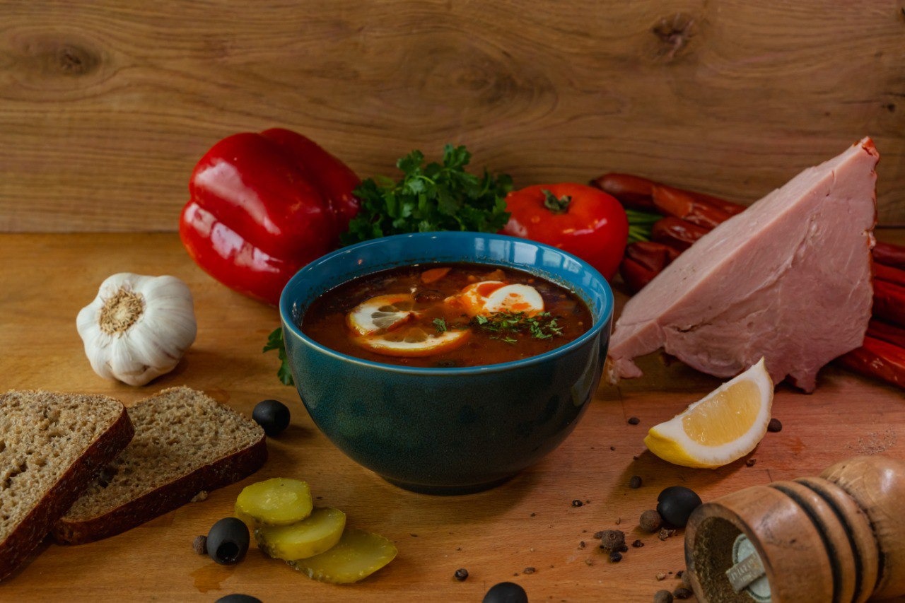 Solyanka w turkusowej miseczce, udekorowana cytryną i jajkiem, na drewnianym tle z chlebem, ogórkami kiszonymi, oliwkami, papryką, pomidorem i wędlinami.