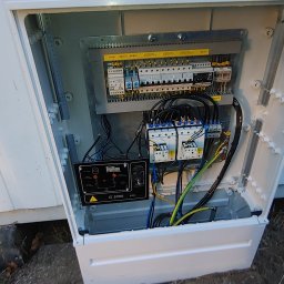 SZR Bydgoszcz - Otwarta skrzynka elektryczna SZR z widocznymi bezpiecznikami, przekaźnikami i okablowaniem, umieszczona na zewnątrz budynku z białym sidingiem.