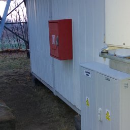 SZR Bydgoszcz - Zewnętrzna szafka SZR z żółtymi znakami ostrzegawczymi na tle białej ściany kontenera, widoczna czerwona skrzynka i konstrukcja wsporcza. Na zewnątrz widok na drzewa bez liści.