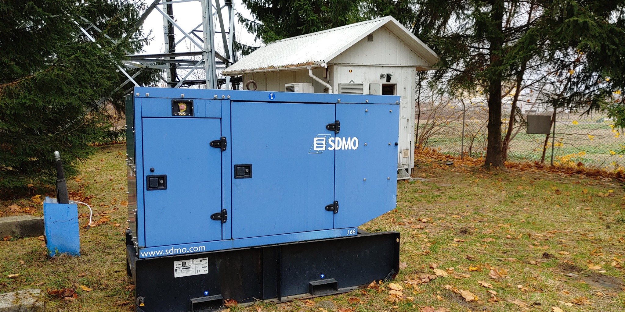 Niebieski agregat prądotwórczy SDMO na zewnątrz, z widocznym budynkiem stacji transformatorowej i wieżą telekomunikacyjną w tle, jesienne liście na trawie.