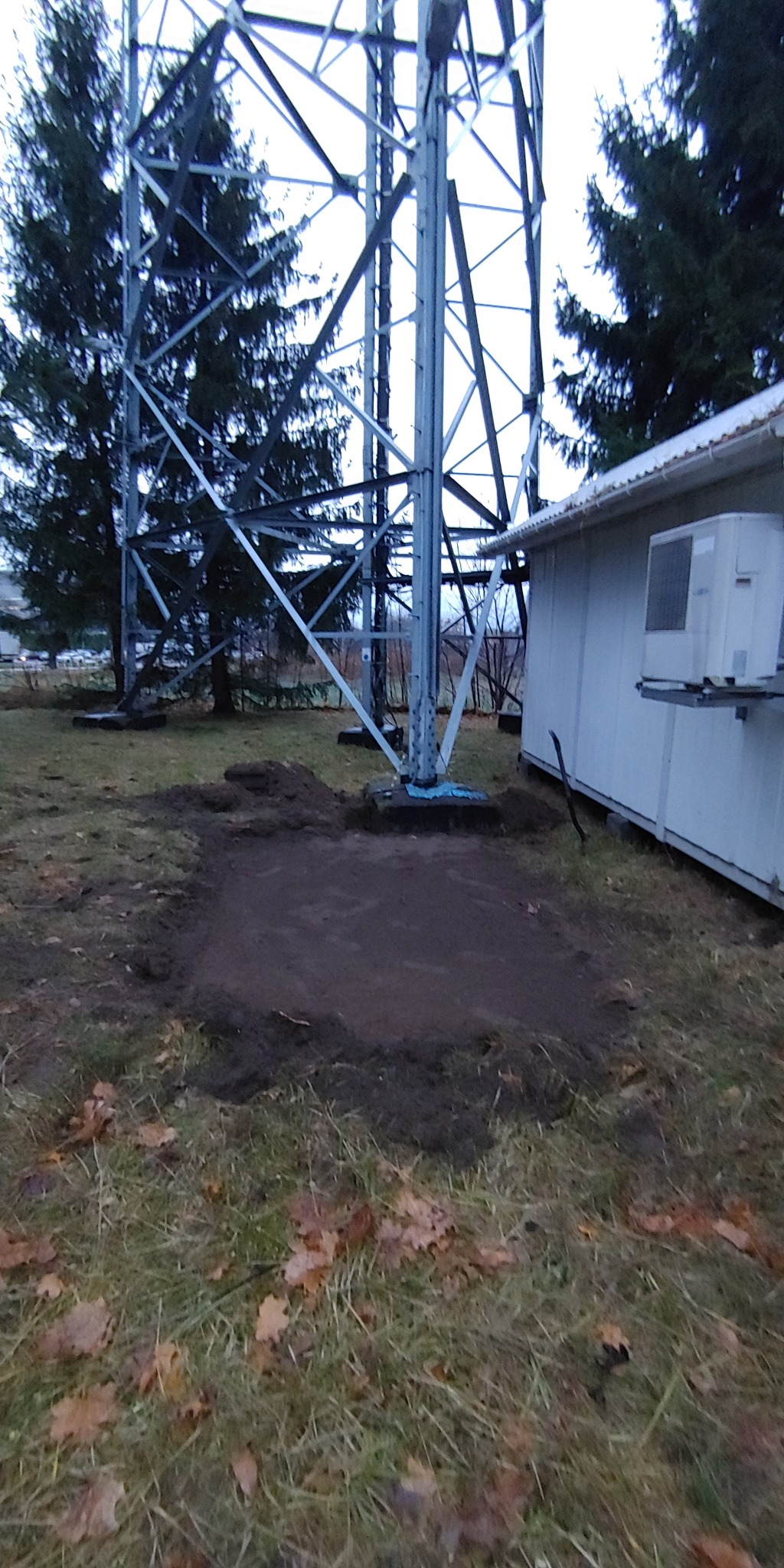 Przygotowanie podłoża pod montaż konstrukcji wsporczej, prawdopodobnie masztu telekomunikacyjnego; widoczny wykop w ziemi, fragment budynku z klimatyzatorem i metalowa konstrukcja masztu.