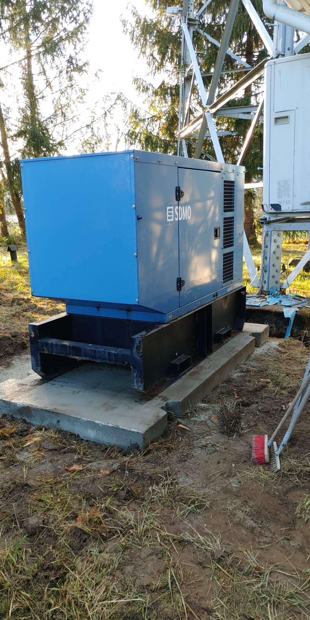 Agregat prądotwórczy SDMO w obudowie na betonowym fundamencie, obok wieża telekomunikacyjna, widoczne ślady prac ziemnych i szczotki do zamiatania.