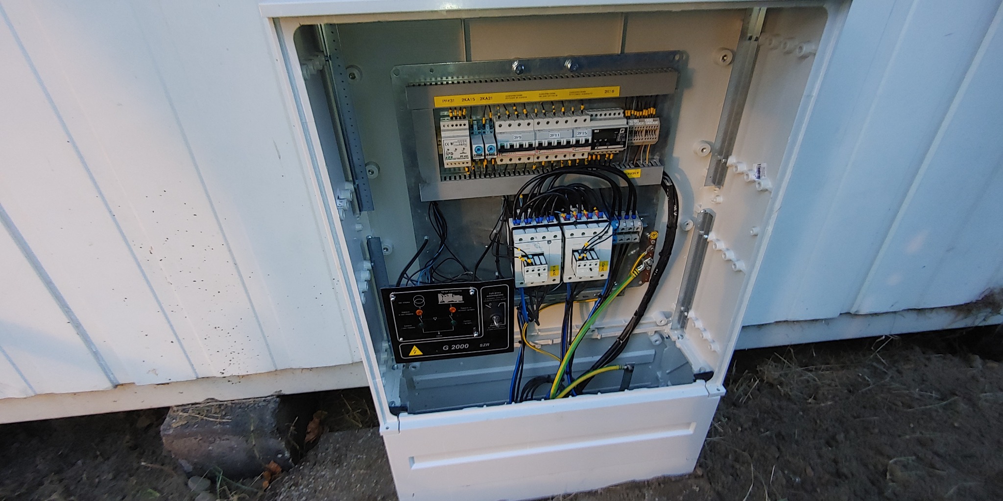 Otwarta skrzynka elektryczna SZR z widocznymi bezpiecznikami, przekaźnikami i okablowaniem, umieszczona na zewnątrz budynku z białym sidingiem.