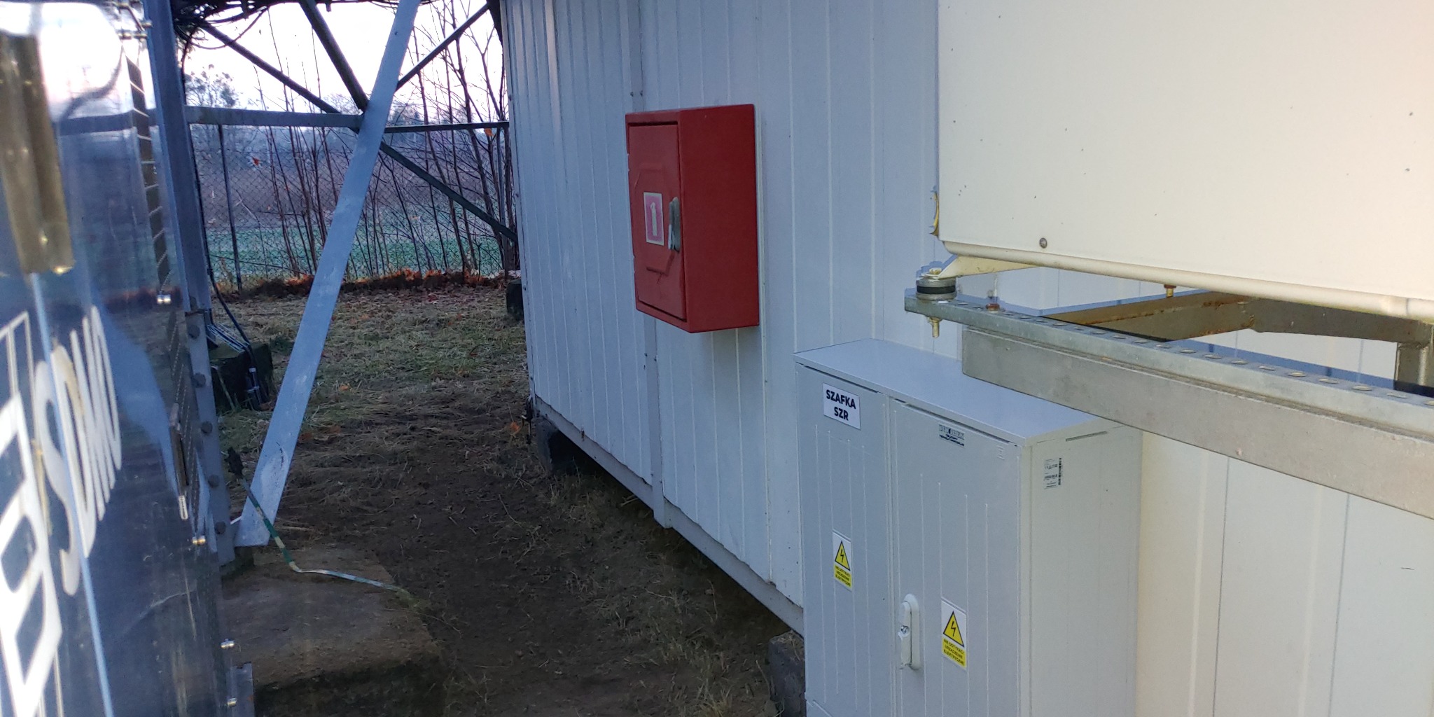 Zewnętrzna szafka SZR z żółtymi znakami ostrzegawczymi na tle białej ściany kontenera, widoczna czerwona skrzynka i konstrukcja wsporcza. Na zewnątrz widok na drzewa bez liści.