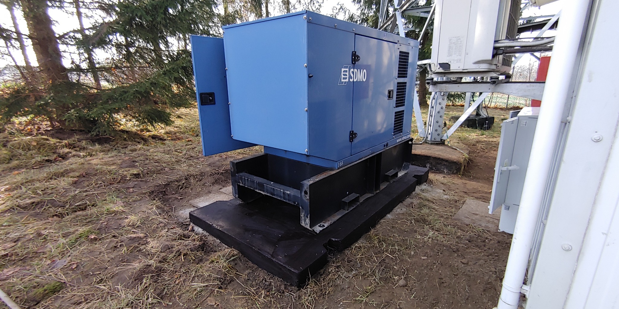 Niebieski agregat prądotwórczy SDMO z otwartymi drzwiczkami, zamontowany na czarnej, betonowej podstawie w pobliżu wieży telekomunikacyjnej na zewnątrz.